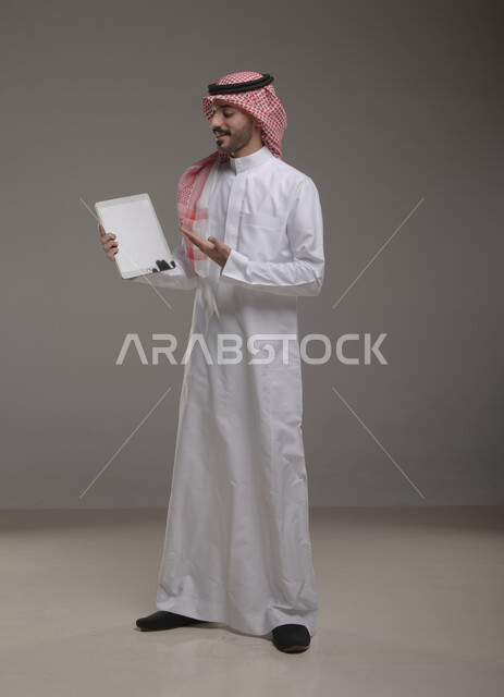 الشرح وتقديم منتجات الشركة , بورتريه لرجل عربي خليجي سعودي يرتدي الزي  التقليدي يحمل بيده  التابليت و يشير بأصبع السبابة اتجاه الجهاز ،التسويق الرقمي و ترويجات إعلانية  ، تطبيقات الأجهزة اللوحية  ، صورة الجسم بالطول الكامل , خلفية رمادية .