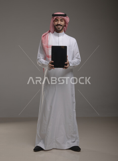 تطبيقات الأجهزة اللوحية  ، بورتريه لرجل عربي خليجي سعودي يرتدي الزي  التقليدي يحمل بيده  التابليت  ،عرض شاشه سوداء فارغة , التسويق الرقمي و ترويجات إعلانية  ، الوقوف باستقامة والنظر إلى الكاميرا , صورة الجسم بالطول الكامل , خلفية رمادية .
