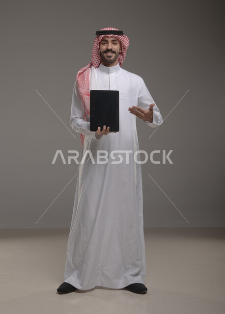 الوقوف باستقامة والنظر إلى الكاميرا , بورتريه لرجل عربي خليجي سعودي يرتدي الزي  التقليدي يحمل بيده  التابليت و يشير بأصبع السبابة اتجاه الجهاز ،عرض شاشه سوداء فارغة , التسويق الرقمي و ترويجات إعلانية  ، تطبيقات الأجهزة اللوحية  ، صورة الجسم بالطول الكامل , خلفية رمادية .