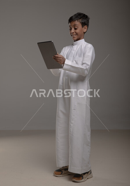 Enjoying watching favorite entertainment programs with gestures of pleasure, portrait of a smiling Saudi Arabian Gulf boy wearing a traditional dress holding a tablet in his hand, using modern technologies to provide generations with useful information, full-length body image, gray background