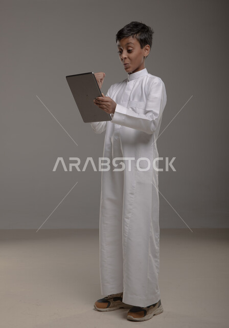 تشجيع المنتخب الوطني بإيماءات الحماس، الاستمتاع في متابعه مباراه كرة القدم، بورتريه لولد عربي خليجي سعودي مبتسم يرتدي الثوب التقليدي يرفع يده بحركات النصر والفوز، استخدام التقنيات الحديثة ، صورة الجسم بالطول الكامل، خلفية رمادية