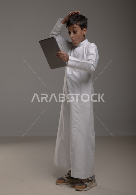Gestures of surprise and astonishment, enjoying watching favorite entertainment programs, portrait of a smiling Gulf Arab Saudi boy wearing a traditional dress holding a tablet in his hand, using modern technologies to provide generations with useful information, full-length body image, gray background