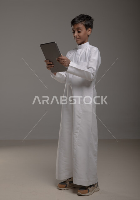 Enjoying watching favorite entertainment programs with gestures of pleasure, portrait of a smiling Saudi Arabian Gulf boy wearing a traditional dress holding a tablet in his hand, using modern technologies to provide generations with useful information, full-length body image, gray background