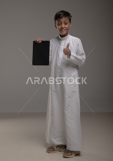 عرض شاشه سوداء فارغه عبر جهاز التابليت، بورتريه لولد عربي خليجي سعودي يرتدي الثوب التقليدي يرفع اصبع الابهام بإيماءات الاعجاب وعلامة الجودة وينظر إلى الكاميرا، استخدام التقنيات الحديثة في تزويد الأجيال بالمعلومات المفيدة، صورة الجسم بالطول الكامل، خلفية رمادية