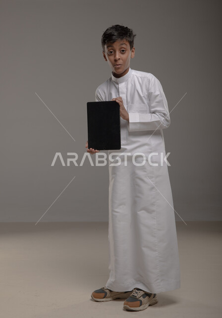 النظر إلى الكاميرا بإيماءات الذهول والتفاجؤ، استخدام التقنيات الحديثة في تزويد الأجيال بالمعلومات المفيدة، بورتريه لولد عربي خليجي سعودي يرتدي الثوب التقليدي يعرض شاشه سوداء فارغه عبر جهاز التابليت، صورة الجسم بالطول الكامل، خلفية رمادية