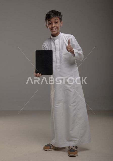 Using modern technologies to provide generations with useful information, displaying a blank black screen on a tablet, a portrait of a Saudi Arabian Gulf boy wearing a traditional dress, raising his thumb with admiration gestures and the quality sign and looking at the camera, full-length body photo, gray background.