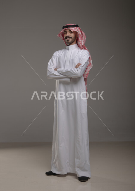  تعابير وجه تدل على السعادة والسرور، بورتريه لشاب عربي خليجي سعودي مبتسم يرتدي الشماغ والثوب التقليدي يقف بأيدي متقاطعة ينظر الى الكاميرا، لغة جسد تدل على الثقة والاعتزاز بالنفس، صورة الجسم بالطول الكامل، خلفية رمادية