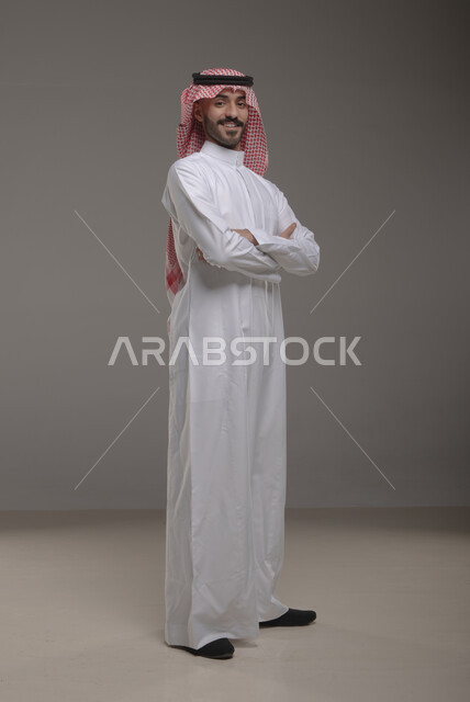 لغة جسد تدل على الثقة والاعتزاز بالنفس، بورتريه لشاب عربي خليجي سعودي مبتسم يرتدي الشماغ والثوب التقليدي يقف بأيدي متقاطعة ينظر الى الكاميرا، ايماءات وجه تدل على السعادة والسرور، صورة الجسم بالطول الكامل، خلفية رمادية