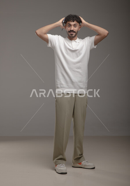  التفاجؤ من شيئ ما، بورتريه لشاب عربي سعودي خليجي يرتدي الزي الكاجوال يضع يديه على رأسه ينظر الى الكاميرا, إيماءات وجه ويدين تدل على الدهشه والصدمه والذهول, تعابير الإستغراب والتعجب, صورة الجسم بالطول الكامل، خلفيه رمادية