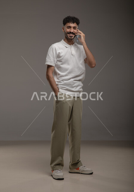 الدردشة والتحدث مع الاصدقاء عبر الجوال، بورتريه لشاب عربي خليجي سعودي مبتسم يرتدي الزي الكاجوال يقوم بالرد على اتصالات الهاتف المحمول ينظر الى الكاميرا، إجراء مكالمة هاتفية، ايماءات السعادة والسرور بسماع اخبار جيدة، صورة الجسم بالطول الكامل، خلفية رمادية