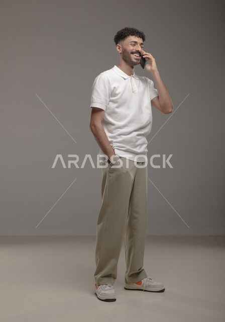 إجراء مكالمة هاتفية، ايماءات السعادة والسرور بسماع اخبار جيدة، الدردشة والتحدث مع الاصدقاء عبر الجوال، بورتريه لشاب عربي خليجي سعودي مبتسم يرتدي الزي الكاجوال يقوم بالرد على اتصالات الهاتف المحمول، صورة الجسم بالطول الكامل، خلفية رمادية