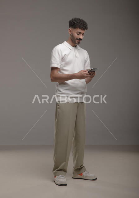 تصفح تطبيقات الهاتف المحمول، الدردشة والتحدث مع الاصدقاء عبر الانترنت، بورتريه لشاب عربي خليجي سعودي مبتسم يرتدي الزي الكاجوال يقوم بمتابعة اخبار وسائل التواصل الاجتماعي باستخدام الجوال، ايماءات الاندماج والتركيز، صورة الجسم بالطول الكامل، خلفية رمادية