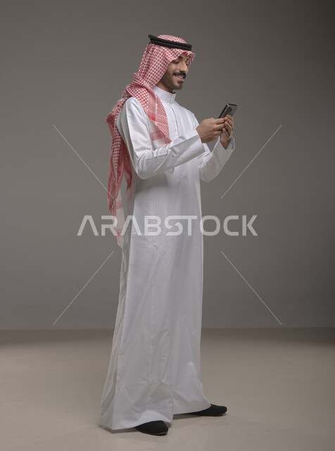 تصفح تطبيقات الهاتف المحمول، الدردشة والتحدث مع الاصدقاء عبر الانترنت، بورتريه لشاب عربي خليجي سعودي مبتسم يرتدي الشماغ والثوب التقليدي يقوم بمتابعة اخبار وسائل التواصل الاجتماعي باستخدام الجوال، ايماءات الاندماج والتركيز، صورة الجسم بالطول الكامل، خلفية رمادية