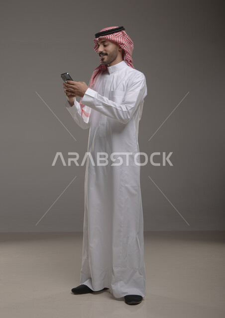 تصفح تطبيقات الهاتف المحمول، الدردشة والتحدث مع الاصدقاء عبر الانترنت، بورتريه لشاب عربي خليجي سعودي مبتسم يرتدي الشماغ والثوب التقليدي يقوم بمتابعة اخبار وسائل التواصل الاجتماعي باستخدام الجوال، ايماءات الاندماج والتركيز، صورة الجسم بالطول الكامل، خلفية رمادية