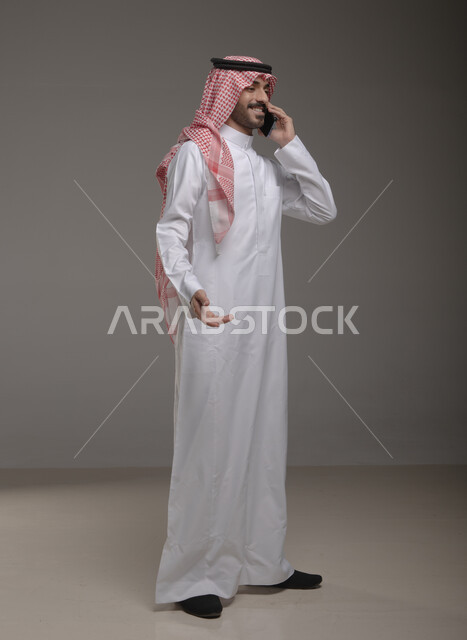 ايماءات السعادة والسرور بسماع اخبار جيدة، إجراء مكالمة هاتفية، الدردشة والتحدث مع الاصدقاء عبر الجوال، بورتريه لشاب عربي خليجي سعودي مبتسم يرتدي الشماغ والثوب التقليدي يقوم بالرد على اتصالات الهاتف المحمول، صورة الجسم بالطول الكامل، خلفية رمادية