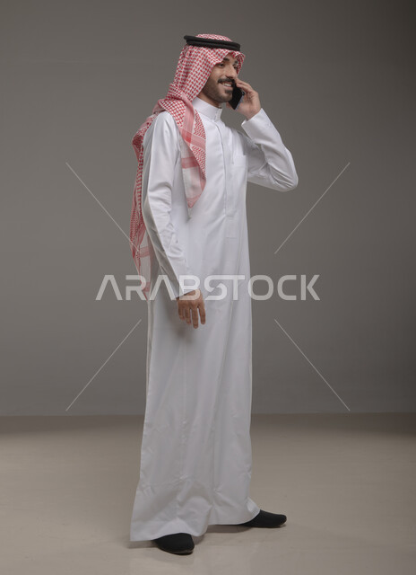 ايماءات السعادة والسرور بسماع اخبار جيدة، إجراء مكالمة هاتفية، الدردشة والتحدث مع الاصدقاء عبر الجوال، بورتريه لشاب عربي خليجي سعودي مبتسم يرتدي الشماغ والثوب التقليدي يقوم بالرد على اتصالات الهاتف المحمول، صورة الجسم بالطول الكامل، خلفية رمادية