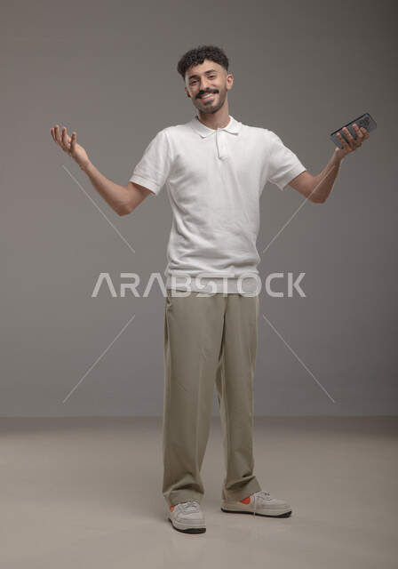تصفح تطبيقات الهاتف المحمول، الدردشة مع الاصدقاء عبر الانترنت، بورتريه لشاب عربي خليجي سعودي مبتسم يرتدي الزي الكاجوال مسرور بمتابعة اخبار وسائل التواصل الاجتماعي باستخدام الجوال ينظر الى الكاميرا، ايماءات الرضا والاعجاب، صورة الجسم بالطول الكامل، خلفية رمادية