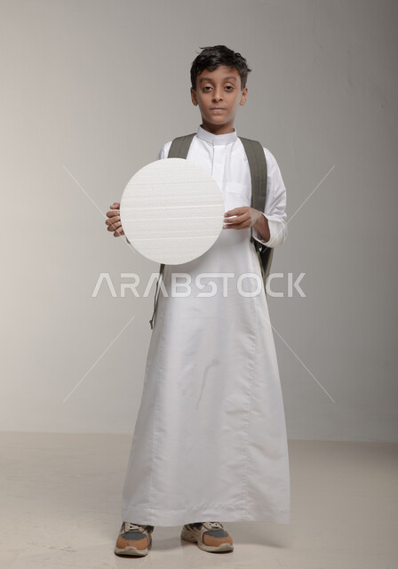 الاعلان عن بداية عام دراسي جديد في المدارس السعودية المتقدمة، الوقوف باستقامة وثقة، موك اب دائري ابيض، بورتريه لفتى عربي خليجي سعودي يرتدي الثوب التقليدي وحقيبة الظهر يحمل بيده لوحة بيضاء مدورة، لوح اعلاني فارغ، صورة الجسم بالطول الكامل، خلفية رمادية