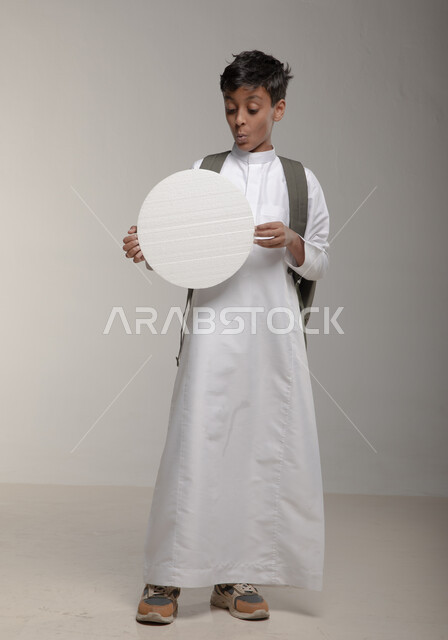 لوح اعلاني فارغ، موك اب دائري ابيض، ايماءات التصفير والدهشة، الاعلان عن بداية عام دراسي جديد في المدارس السعودية المتقدمة، بورتريه لفتى عربي خليجي سعودي يرتدي الثوب التقليدي وحقيبة الظهر يحمل بيده لوحة بيضاء مدورة، صورة الجسم بالطول الكامل، خلفية رمادية