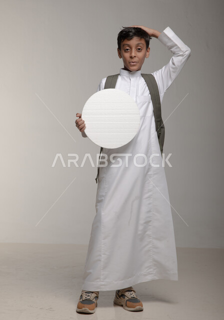 موك اب دائري ابيض، ايماءات الاعجاب والدهشة، الاعلان عن بداية عام دراسي جديد في المدارس السعودية المتقدمة، بورتريه لفتى عربي خليجي سعودي يرتدي الثوب التقليدي وحقيبة الظهر يحمل بيده لوحة بيضاء مدورة، لوح اعلاني فارغ، صورة الجسم بالطول الكامل، خلفية رمادية