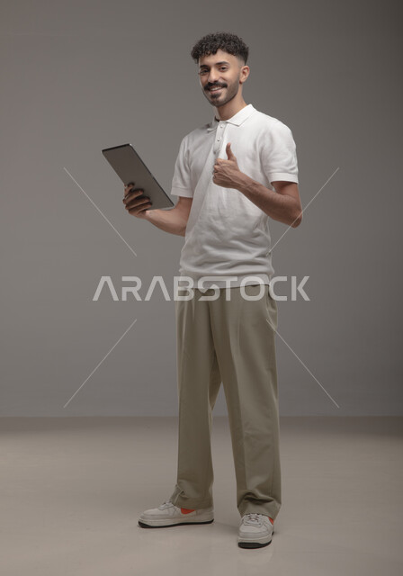 Raising a thumb to indicate satisfaction and admiration for quality, using advanced technology for electronic digital marketing, displaying tablet applications and services, portrait of a smiling Saudi Arabian Gulf man wearing a casual outfit holding a tablet in his hand, full-length body portrait, gray background