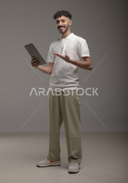 Using advanced technology for electronic digital marketing, displaying tablet applications and services, portrait of a smiling Saudi Arabian Gulf man wearing a casual outfit holding a tablet in his hand and pointing at it, full-length body portrait, gray background
