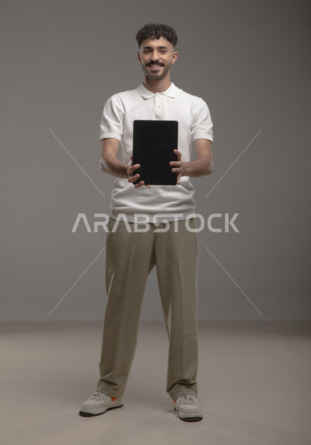 Using advanced technology for electronic digital marketing, displaying tablet applications and services, portrait of a smiling Saudi Arabian Gulf man wearing a casual outfit holding a tablet with a blank black screen in his hands, full-length body portrait, gray background