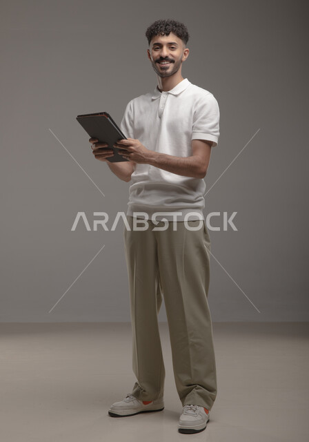 تصفح تطبيقات جهاز التابلت، الدردشة والتحدث مع الاصدقاء عبر الانترنت، بورتريه لشاب عربي خليجي سعودي مبتسم يرتدي الزي الكاجوال يقوم بمتابعة اخبار وسائل التواصل الاجتماعي باستخدام التابلت، النظر الى الكاميرا بايماءات السعادة والسرور، صورة الجسم بالطول الكامل، خلفية رمادية
