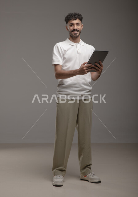 تصفح تطبيقات جهاز التابلت، الدردشة والتحدث مع الاصدقاء عبر الانترنت، بورتريه لشاب عربي خليجي سعودي مبتسم يرتدي الزي الكاجوال يقوم بمتابعة اخبار وسائل التواصل الاجتماعي باستخدام التابلت، النظر الى الكاميرا بايماءات السعادة والسرور، صورة الجسم بالطول الكامل، خلفية رمادية