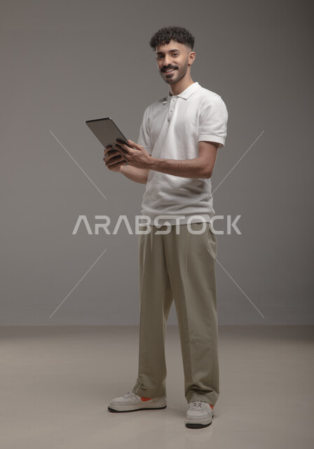 تصفح تطبيقات جهاز التابلت، الدردشة والتحدث مع الاصدقاء عبر الانترنت، بورتريه لشاب عربي خليجي سعودي مبتسم يرتدي الزي الكاجوال يقوم بمتابعة اخبار وسائل التواصل الاجتماعي باستخدام التابلت، النظر الى الكاميرا بايماءات السعادة والسرور، صورة الجسم بالطول الكامل، خلفية رمادية