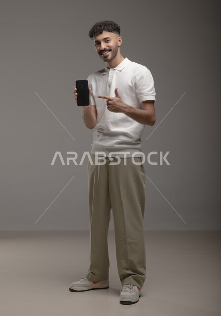 Using advanced technology for electronic digital marketing, displaying mobile phone applications and services, portrait of a smiling Saudi Gulf Arab young man wearing a casual outfit holding a mobile phone with a blank black screen in his hand and pointing at it with his index finger, full-length body portrait, gray background