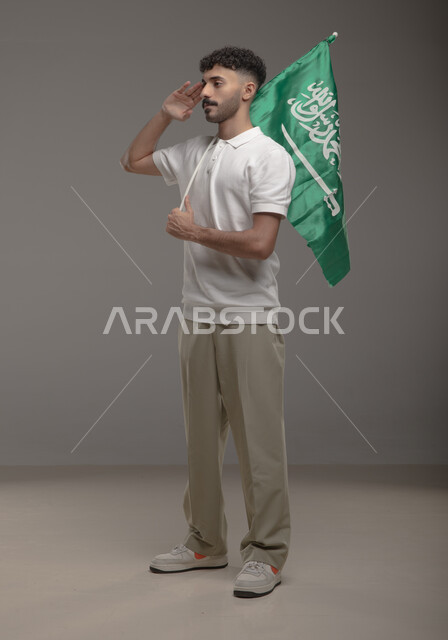 Proudly saluting the country and taking pride in the national identity, Saudi National Day, September 23, Portrait of a smiling Gulf Arab Saudi man wearing casual clothes, holding the flag of the Kingdom of Saudi Arabia in his hand and looking at something, Flag Day, March 11, full-length body portrait, gray background