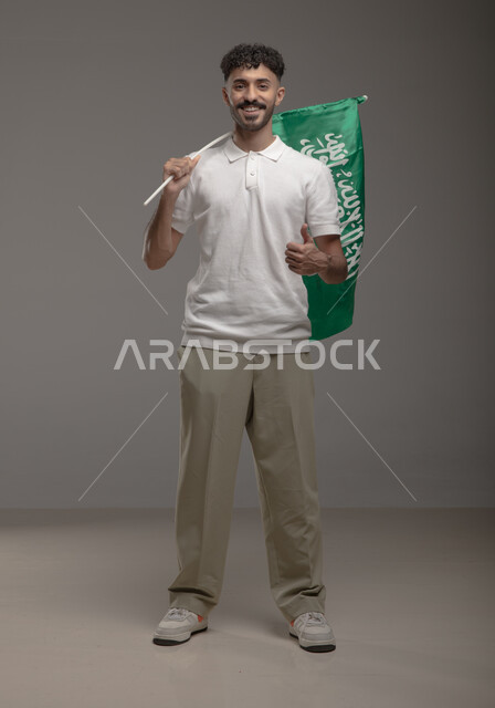 اليوم الوطني السعودي 23 سبتمبر، رفع اصبع الابهام بتعبيرات الإعجاب بالهوية الوطنية، بورتريه لشاب عربي خليجي سعودي مبتسم يرتدي الزي الكاجوال يحمل بيده علم المملكة العربيه السعودية وينظر إلى الكاميرا، يوم العلم 11 مارس، صورة الجسم بالطول الكامل، خلفية رمادية
