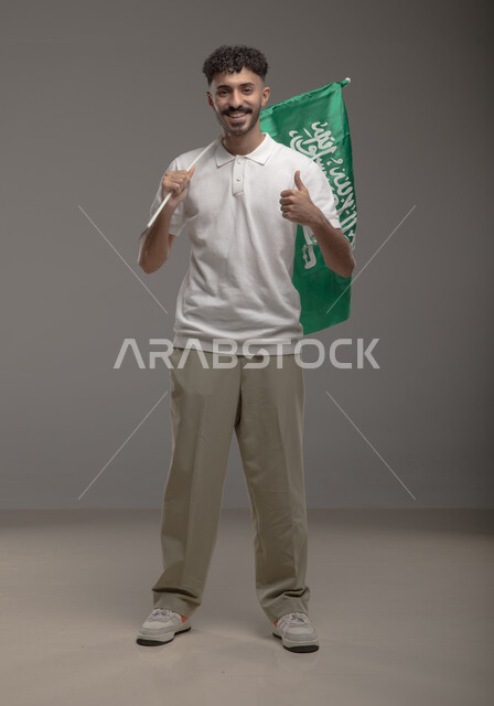 رفع اصبع الابهام بتعبيرات الإعجاب بالهوية الوطنية، بورتريه لشاب عربي خليجي سعودي مبتسم يرتدي الزي الكاجوال يحمل بيده علم المملكة العربيه السعودية وينظر إلى الكاميرا، يوم العلم 11 مارس، اليوم الوطني السعودي 23 سبتمبر، صورة الجسم بالطول الكامل، خلفية رمادية