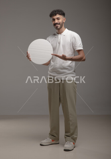 النظر الى الكاميرا بايماءات الإعجاب والسرور، لوح إعلاني فارغ، موك اب دائري أبيض، بورتريه لرجل عربي خليجي سعودي مبتسم يرتدي الزي الكاجوال و يشير بيده إلى لوحه بيضاء مدوره، الوقوف باستقامه وثقه، صورة الجسم بالطول الكامل، خلفية رماديه