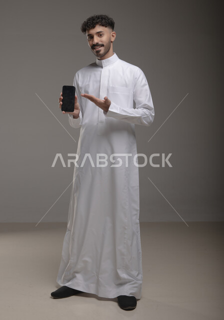 Using advanced technology for electronic digital marketing, pointing to a blank black screen on a mobile phone, displaying modern applications and services, a portrait of a smiling Gulf Arab Saudi man wearing a traditional thobe and holding a mobile phone in his hand, full-length body photo, gray background