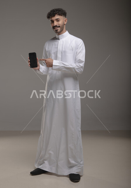 الإشارة باصبع السبابه إلى شاشة سوداء فارغة عبر الهاتف المحمول ،استخدام التكنولوجيا المتطورة للتسويق الرقمي الالكتروني، عرض التطبيقات الحديثة، بورتريه لشاب عربي خليجي سعودي مبتسم يرتدي الثوب التقليدي و يحمل بيده جوال، صورة الجسم بالطول الكامل، خلفية رمادية