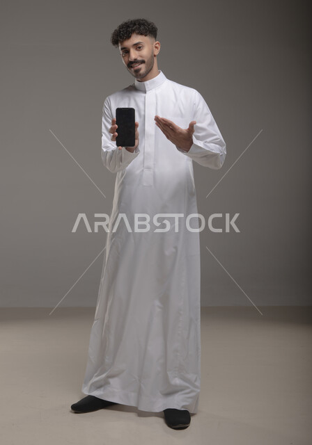 Pointing to a blank black screen on a mobile phone, displaying modern applications and services, a portrait of a smiling Saudi Gulf Arab young man wearing a traditional dress and holding a mobile phone in his hand, using advanced technology for electronic digital marketing, full-length body image, gray background