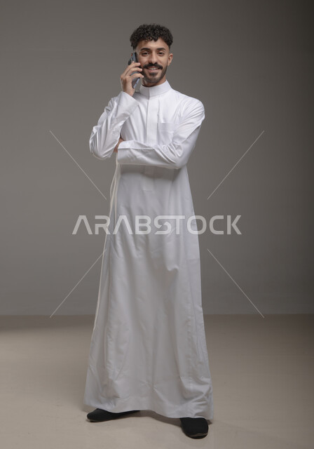 الدردشة والتحدث مع الاقارب عبر الجوال، ايماءات السعادة والسرور بسماع اخبار جيدة، الرد على اتصالات الهاتف المحمول، بورتريه لشاب عربي خليجي سعودي مبتسم يرتدي الثوب التقليدي يقوم بإجراء مكالمة هاتفية وينظر إلى الكاميرا، صورة الجسم بالطول الكامل، خلفية رمادية
