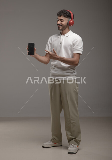 عرض تطبيقات الاغاني والتسجيلات الصوتية في الهاتف المحمول، الاستماع إلى الأغاني عبر الهيدفون، بورتريه لشاب عربي خليجي سعودي يرتدي الزي الكاجوال وسماعة الرأس يحمل بيده جوال بشاشة سوداء فارغة ويشير اليه، صورة الجسم بالطول الكامل، خلفية رمادية