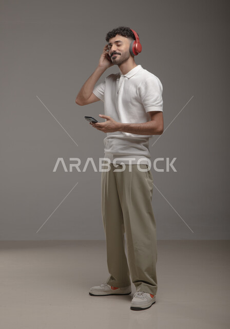 ايماءات الاندماج والاستمتاع، عرض تطبيقات الاغاني والتسجيلات الصوتية في الهاتف المحمول، الاستماع إلى الأغاني عبر الهيدفون، بورتريه لشاب عربي خليجي سعودي يرتدي الزي الكاجوال وسماعة الرأس يحمل بيده جوال، صورة الجسم بالطول الكامل، خلفية رمادية