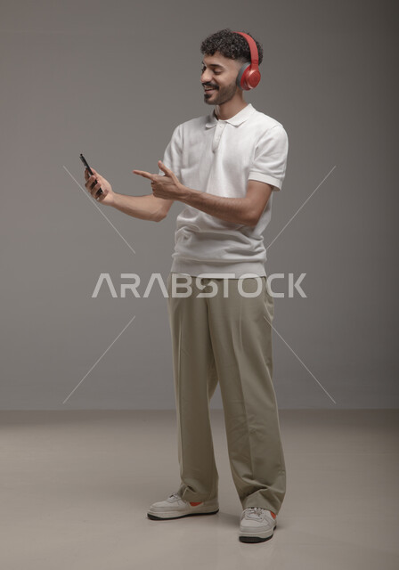 عرض تطبيقات الاغاني والتسجيلات الصوتية في الهاتف المحمول، الاستماع إلى الأغاني عبر الهيدفون، بورتريه لشاب عربي خليجي سعودي يرتدي الزي الكاجوال وسماعة الرأس يحمل بيده جوال ويشير اليه باصبع السبابة، صورة الجسم بالطول الكامل، خلفية رمادية
