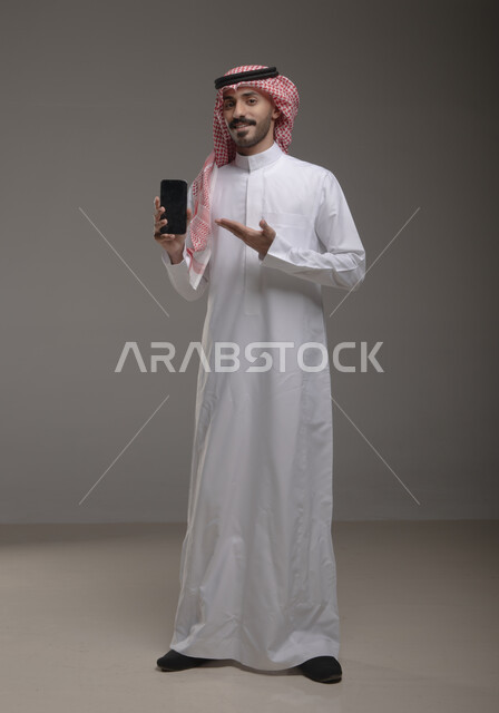 مفهوم التسويق الرقمي الالكتروني، عرض تطبيقات وخدمات الهاتف المحمول، بورتريه لشاب عربي خليجي سعودي مبتسم يرتدي االشماغ ولثوب التقليدي يحمل بيده جوال بشاشة سوداء فارغة ويشير اليه، استخدام التكنولوجيا المتطورة، صورة الجسم بالطول الكامل، خلفية رمادية