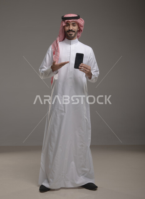 مفهوم التسويق الرقمي الالكتروني، عرض تطبيقات وخدمات الهاتف المحمول، بورتريه لشاب عربي خليجي سعودي مبتسم يرتدي االشماغ ولثوب التقليدي يحمل بيده جوال بشاشة سوداء فارغة ويشير اليه، استخدام التكنولوجيا المتطورة، صورة الجسم بالطول الكامل، خلفية رمادية