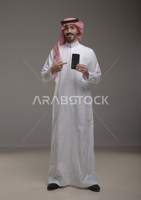 Using advanced technology for electronic digital marketing, displaying mobile phone applications and services, a portrait of a smiling Saudi Gulf Arab young man wearing a shemagh and traditional thobe, holding a mobile phone with a blank black screen in his hand and pointing at it with his index finger, full-length body portrait, gray background.