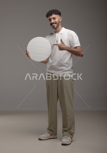 النظر الى الكاميرا بإيماءات الإعجاب والسرور، لوح إعلاني فارغ، موك اب دائري أبيض، بورتريه لرجل عربي خليجي سعودي مبتسم يرتدي الزي الكاجوال و يشير باصبع السبابه إلى لوحه بيضاء مدوره، الوقوف باستقامه وثقه، صورة الجسم بالطول الكامل، خلفية رماديه