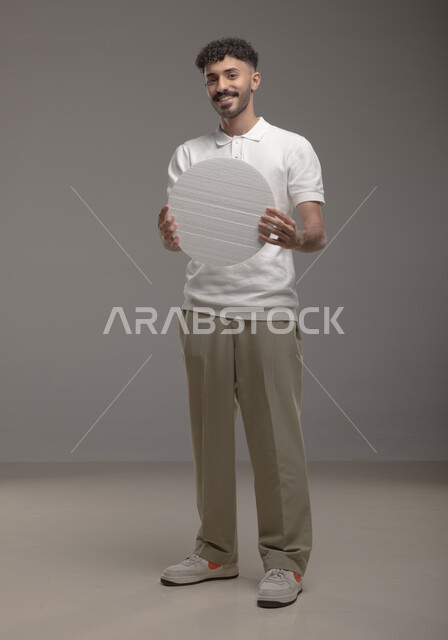 صورة الجسم بالطول الكامل، لوح إعلاني فارغ، موك اب دائري أبيض، بورتريه لرجل عربي خليجي سعودي مبتسم يرتدي الزي الكاجوال و يحمل بيده لوحه بيضاء مدوره، الوقوف باستقامه وثقه، النظر الى الكاميرا بايماءات الإعجاب والسرور، خلفية رماديه