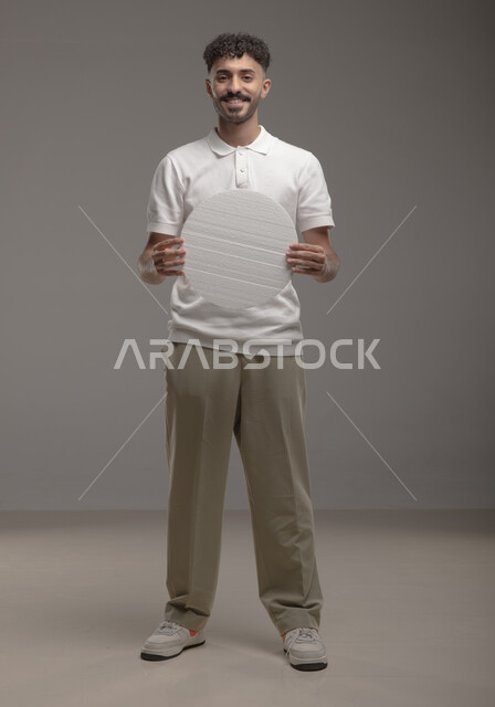 صورة الجسم بالطول الكامل، لوح إعلاني فارغ، موك اب دائري أبيض، بورتريه لرجل عربي خليجي سعودي مبتسم يرتدي الزي الكاجوال و يحمل بيده لوحه بيضاء مدوره، الوقوف باستقامه وثقه، النظر الى الكاميرا بايماءات الإعجاب والسرور، خلفية رماديه