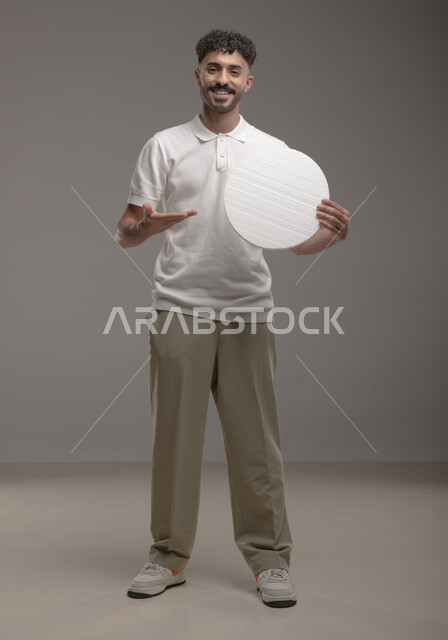 موك اب دائري أبيض، النظر الى الكاميرا بإيماءات السرور والاعجاب، لوح إعلاني فارغ، بورتريه لرجل عربي خليجي سعودي مبتسم يرتدي الزي الكاجوال و يشير بيده إلى لوحه بيضاء مدوره، الوقوف باستقامه وثقه، صورة الجسم بالطول الكامل، خلفية رماديه