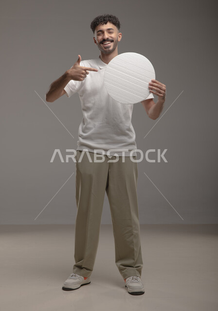 النظر الى الكاميرا بإيماءات السرور والاعجاب، لوح إعلاني فارغ، موك اب دائري أبيض، بورتريه لرجل عربي خليجي سعودي مبتسم يرتدي الزي الكاجوال و يشير باصبع السبابه إلى لوحه بيضاء مدوره، الوقوف باستقامه وثقه، صورة الجسم بالطول الكامل، خلفية رماديه